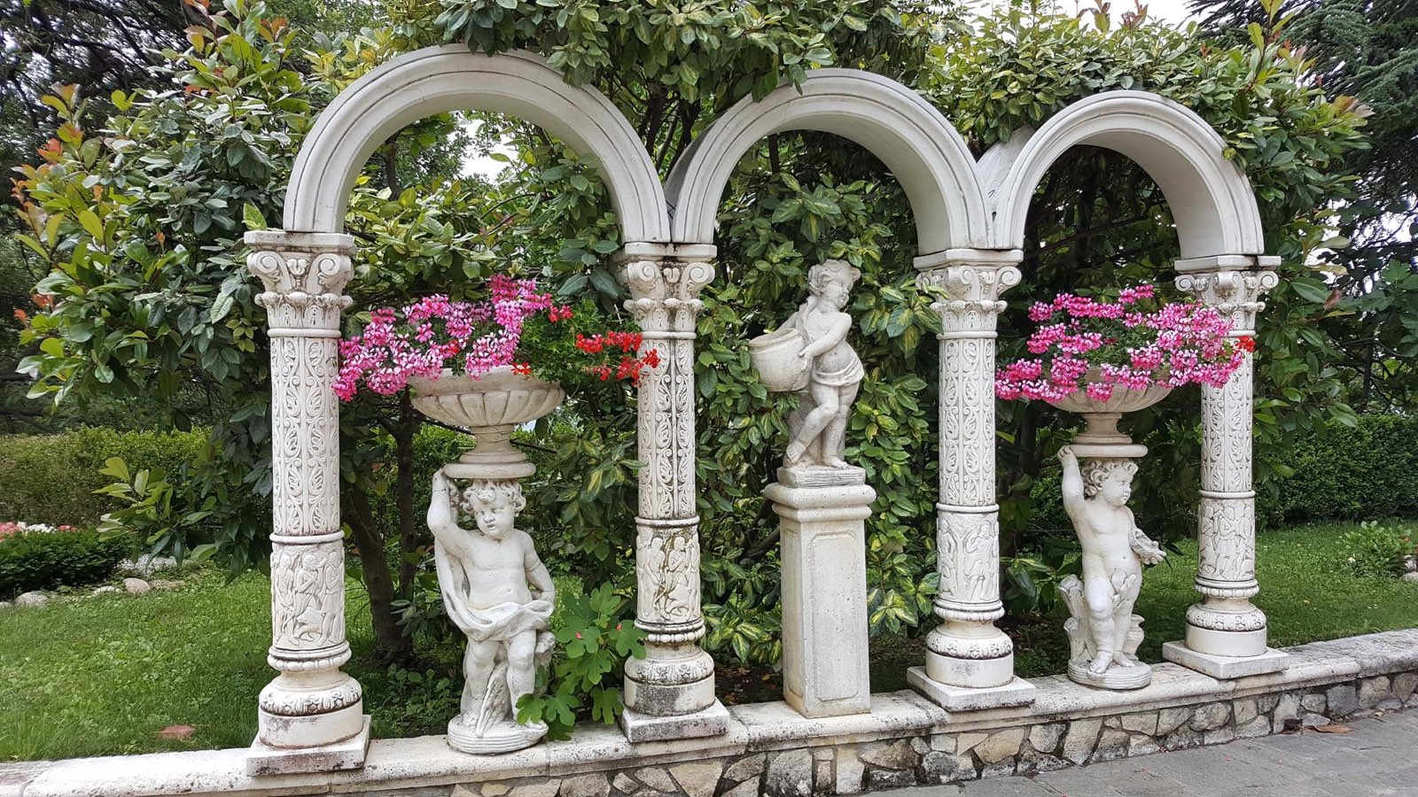 Tre archi bianchi con colonne decorate, statue di putti e vasi di gerani rosa