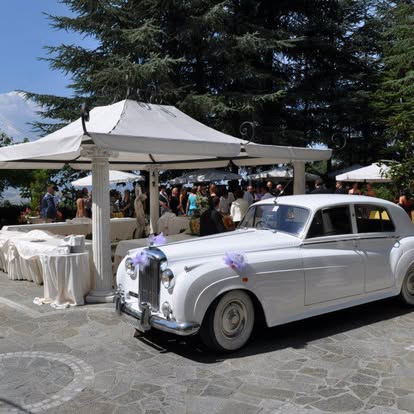 Un'auto d'epoca bianca con nastri lilla davanti al gazebo, con gli ospiti in giardino