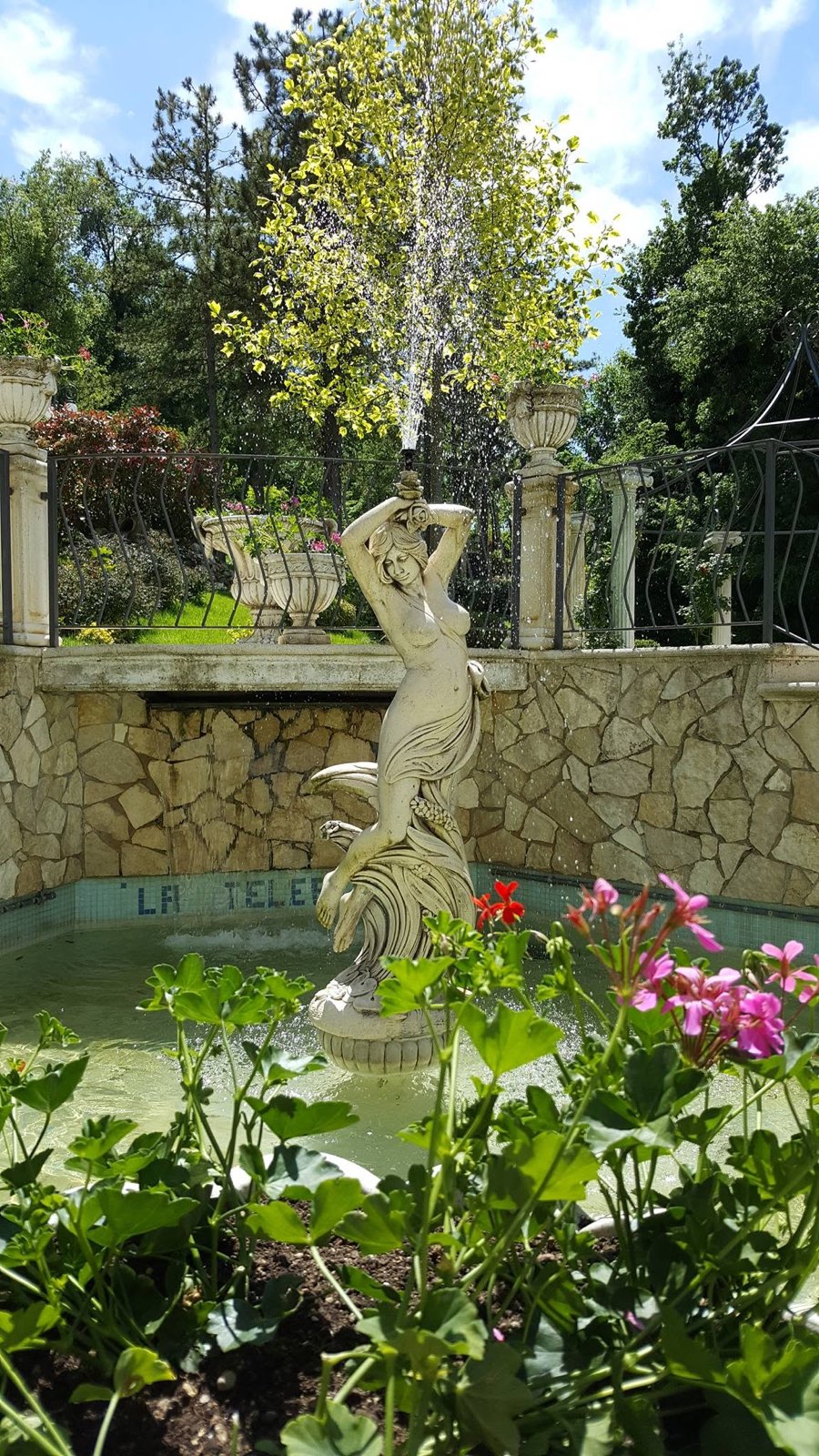 Statua femminile al centro di una fontana, con gerani in primo piano