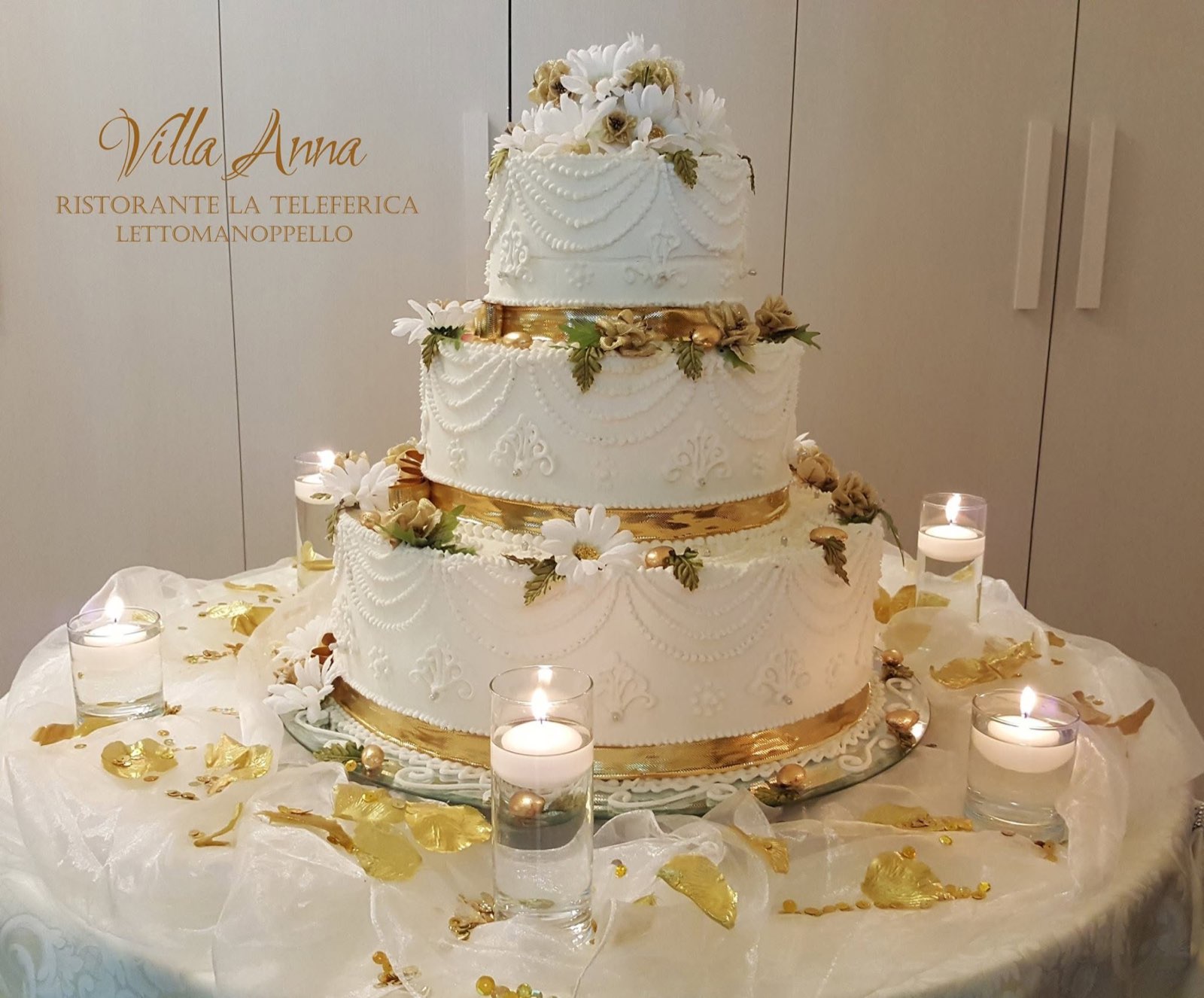 Torta a tre piani bianca con nastri dorati, margherite e candele galleggianti
